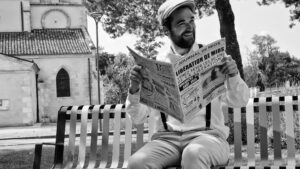 un homme assis sur un banc qui tient un journal devant l'église de Mios