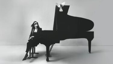 jeune femme assise devant un piano habillée en noir, elle porte des lunettes. Image en noir et blanc.