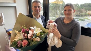 femme habillée en gris qui tient un bébé dans les bras, un homme qui tient un bouquet de fleurs est à côté d'elle