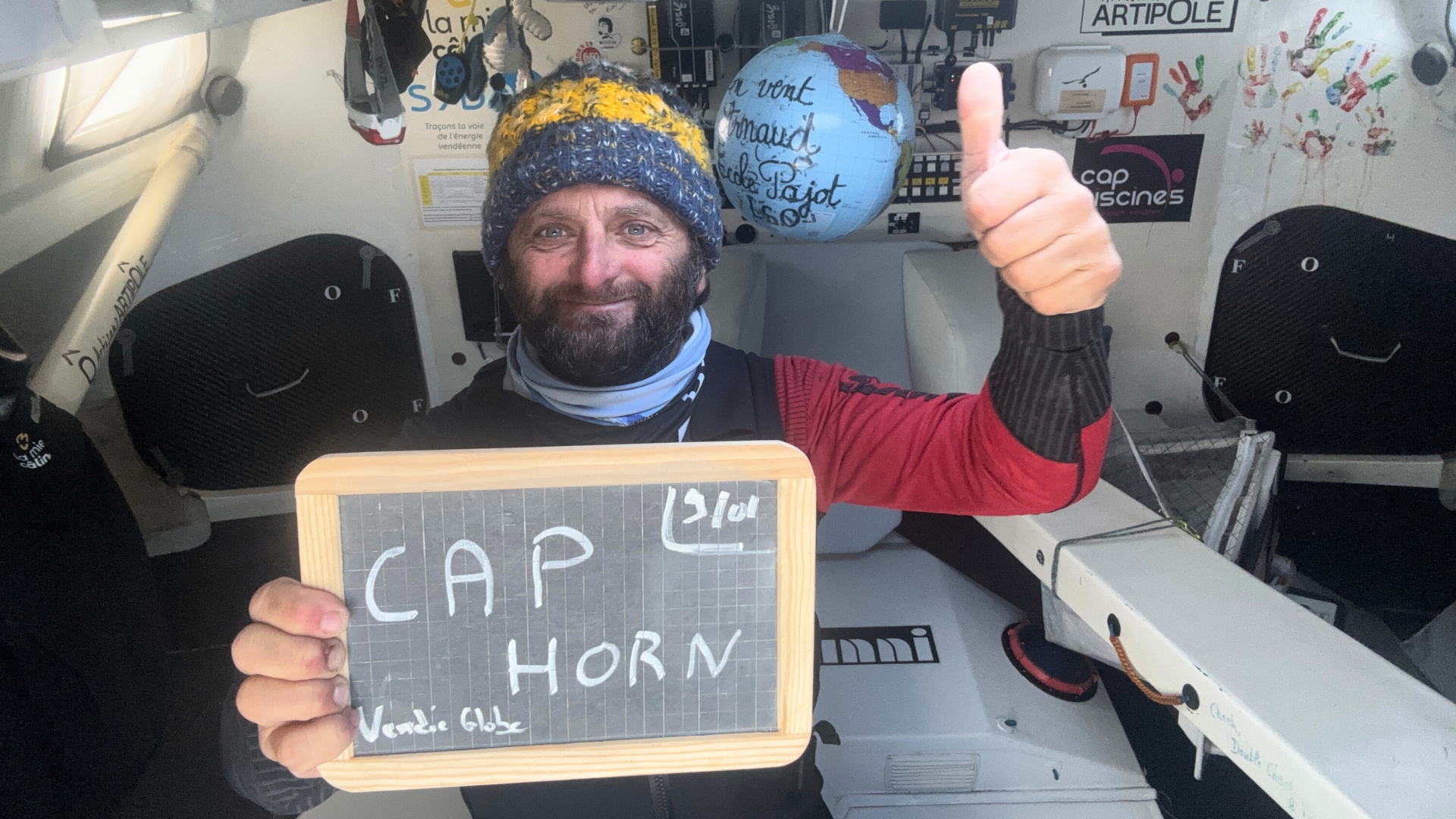 Homme barbu qui porte un bonnet dans la cabine d'un voilier, il tient une ardoise avec la mention "Cap Horn"