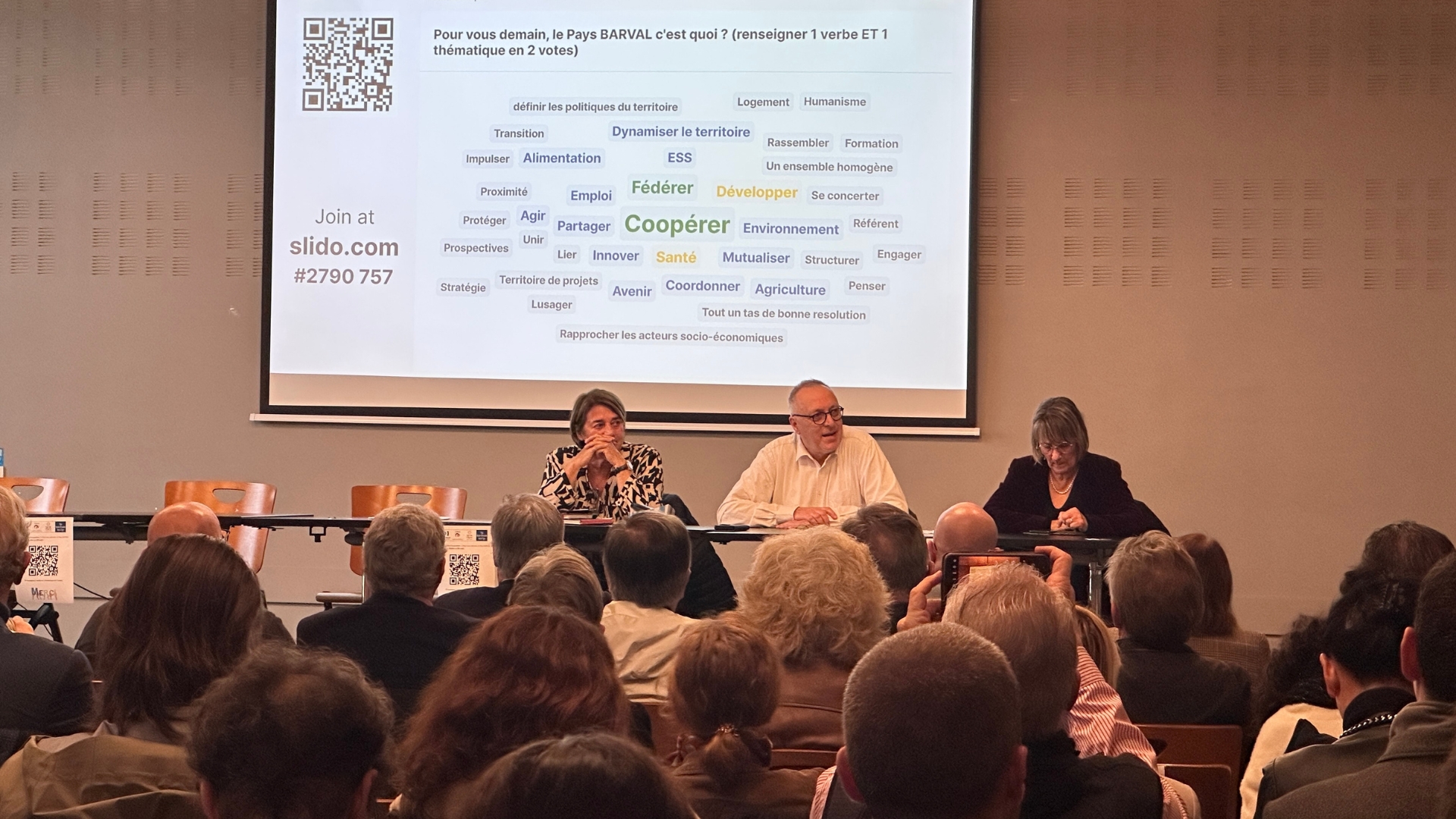 3 personnes sur une scène assises derrière des tables. le public face à elles au premier plan