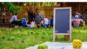 Groupe d'enfants au second plan qui écoutent une animatrice de loisir. Au premier plan, il y a une ardoise sur pieds et un jeu de l'oie grandeur nature