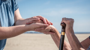 Main d'une aide soignante qui tient la main d'une personne âgée, qui tient également sa canne de l'autre main. On aperçoit la main en fond