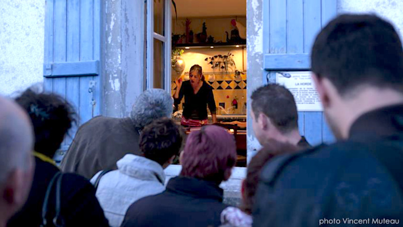 Un groupe de personnes regardent une dame dans sa cuisine à traver sla fenêtre