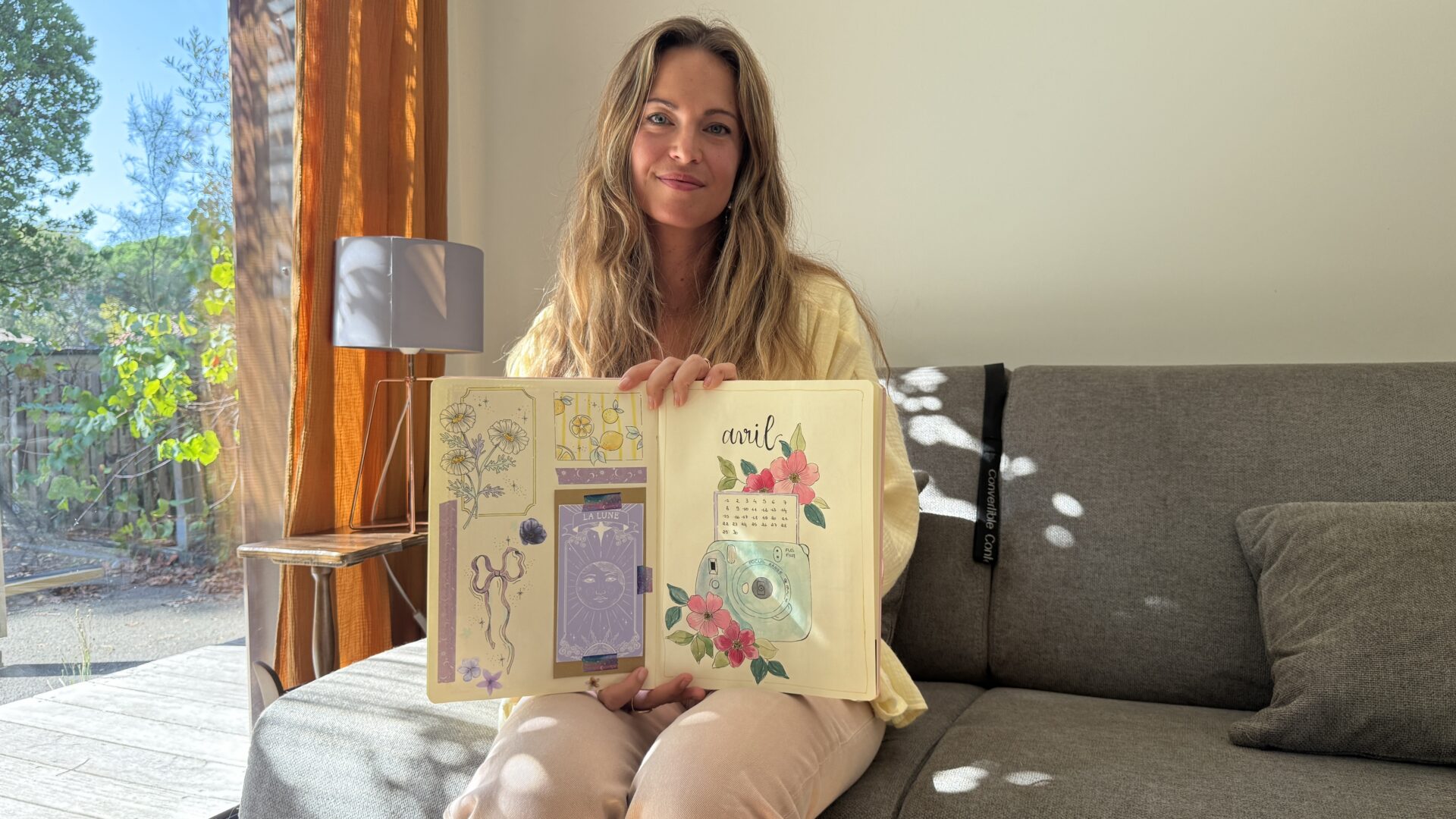 Une femme assise sur un canapé gris tient dans ses mains un carnet décoré de dessins et collages.