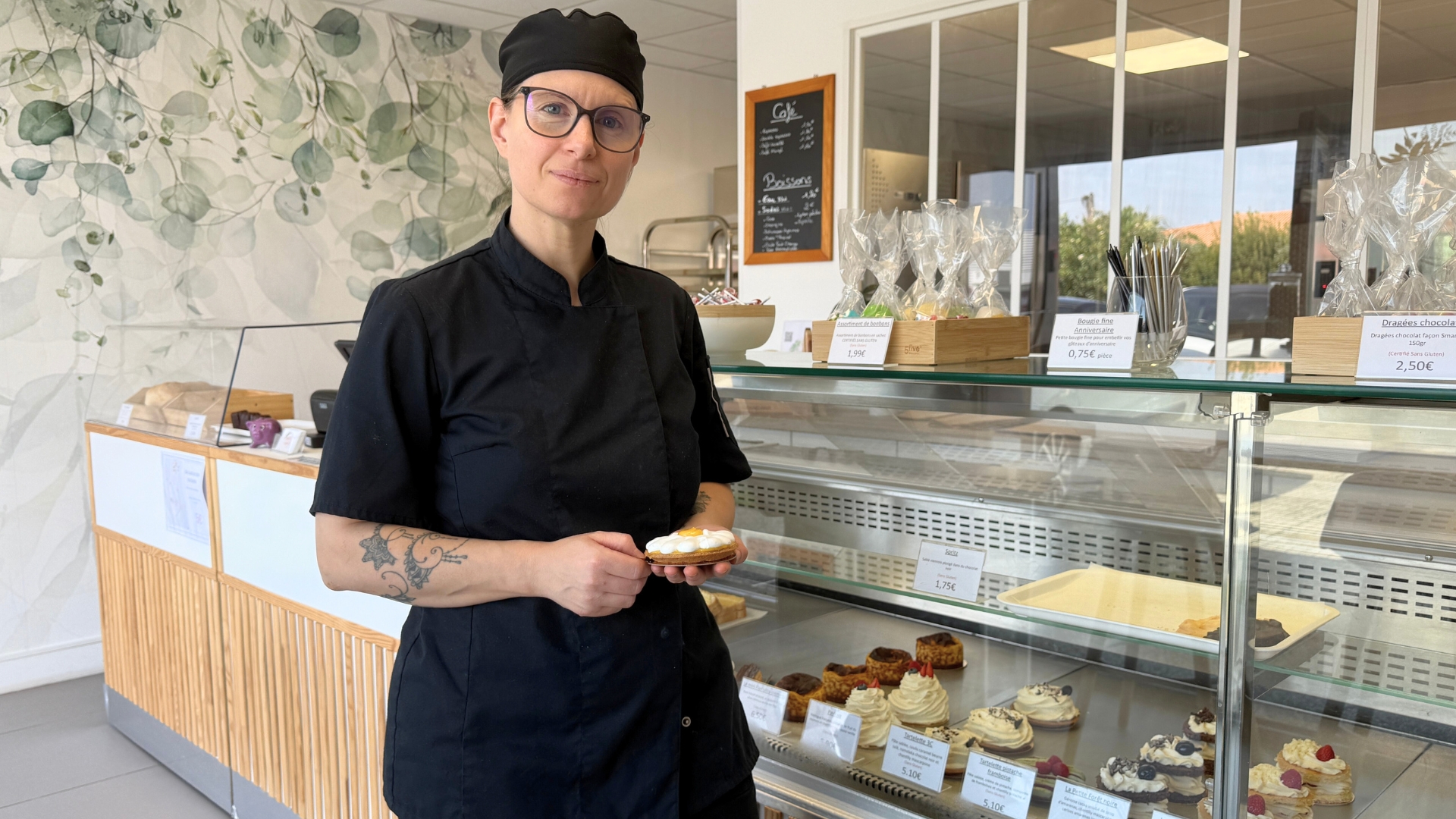 femme souriante habillée en noir qui tient une patisserie dans les mains. Elle pose devant une vitrine de pâtisseries