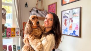 jeune femme souriante qui porte un cocker roux dans les bras, en arrière plan : des tableaux au mur et un sac