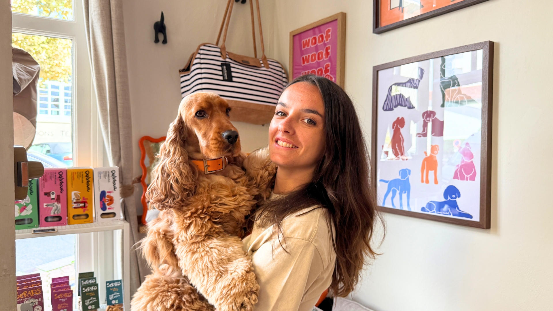 jeune femme souriante qui porte un cocker roux dans les bras, en arrière plan : des tableaux au mur et un sac