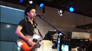 Un jeune homme avec un tee shirt noirjoue de la guitare et chante sur scène