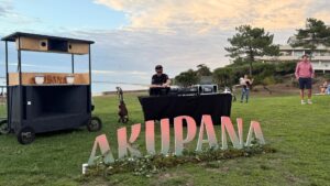 dj derrière une table de mixage avec le bassin d'arcachon en arrière plan et des grosses lettres formant le mot "AKUPANA" au premier plan