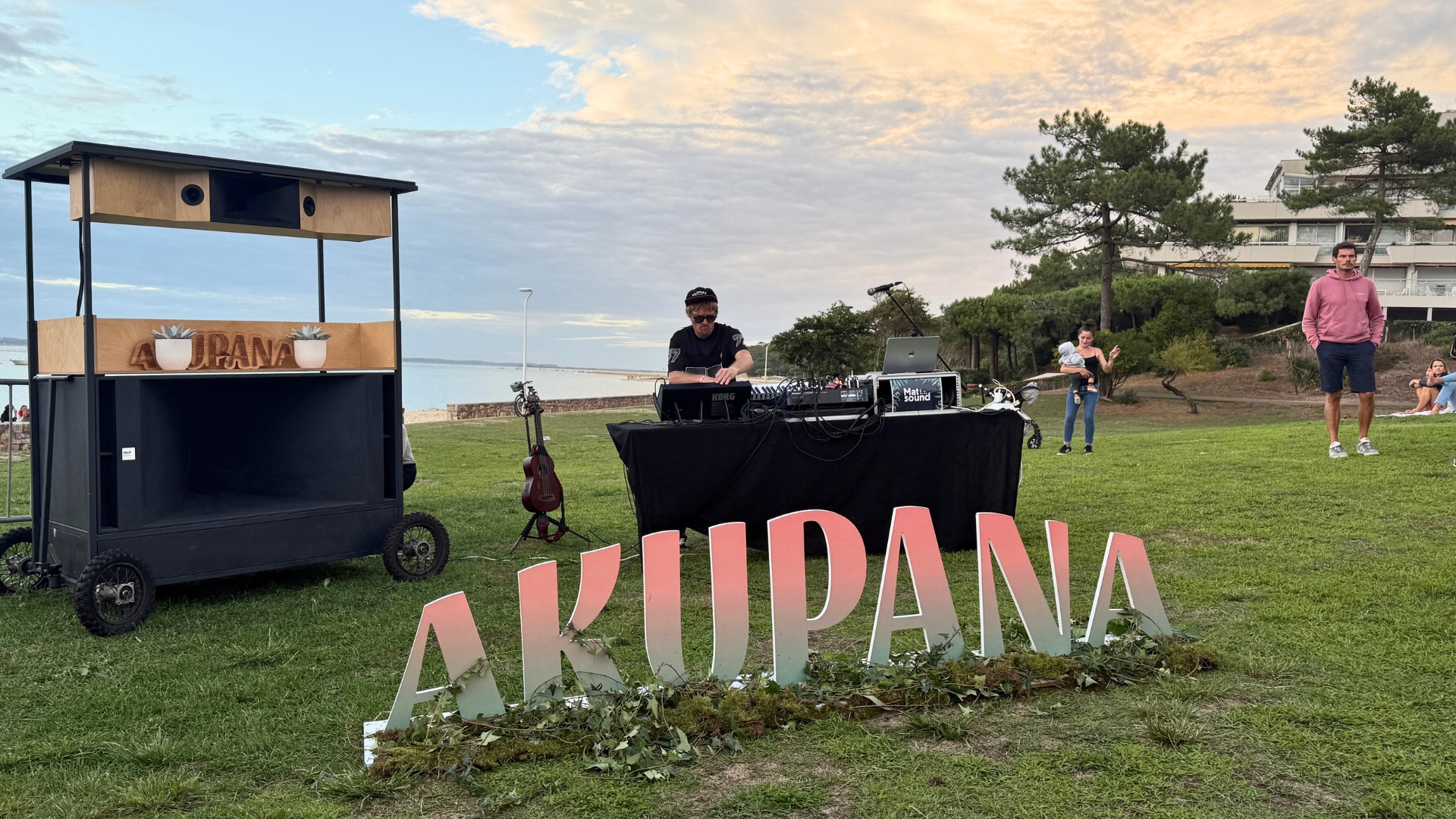 dj derrière une table de mixage avec le bassin d'arcachon en arrière plan et des grosses lettres formant le mot "AKUPANA" au premier plan