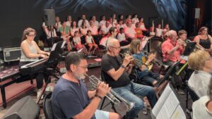 dans une salle noire, des personnes jouent de la musique avec différents instruments. En arrière plan, des choristes sont habillés en blanc et constituent la chorale de la ville d'andernos