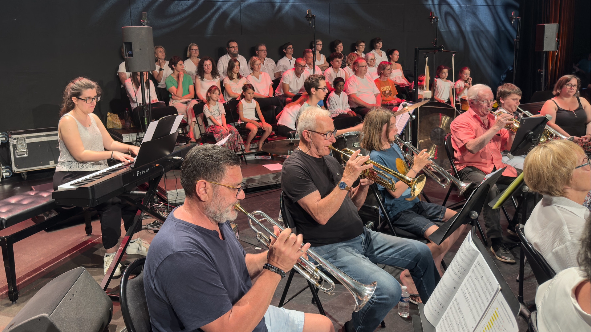 dans une salle noire, des personnes jouent de la musique avec différents instruments. En arrière plan, des choristes sont habillés en blanc et constituent la chorale de la ville d'andernos