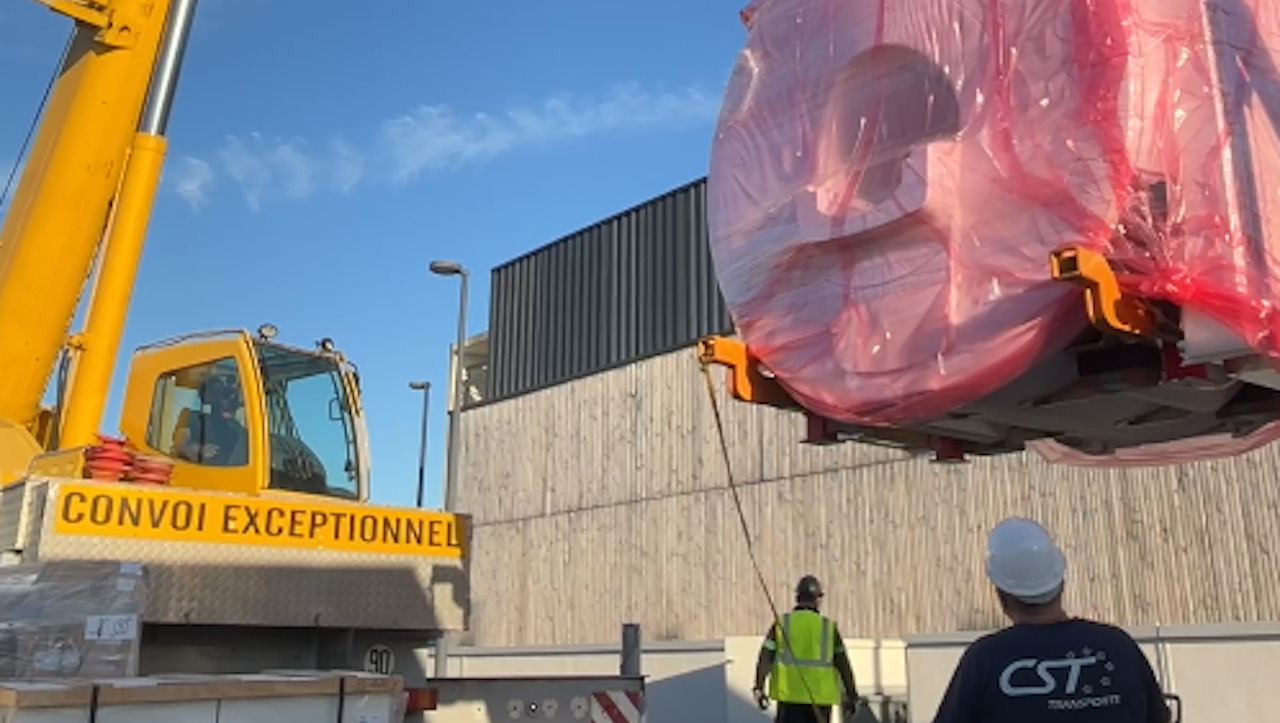 Une grue porte un gros colis emballée dans une bache rose devant un batiment en bois.