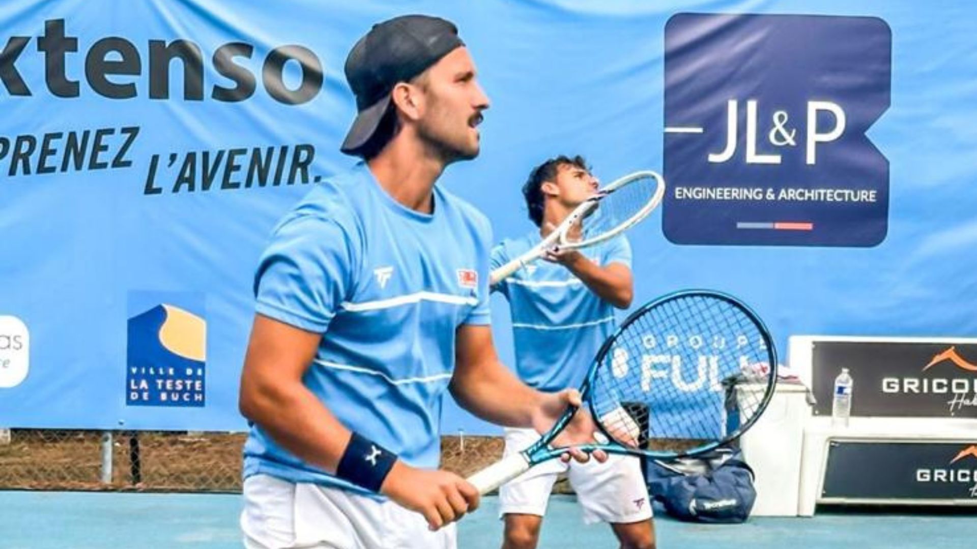 Deux hommes en teeshirt bleu clair tienent des raquettes et joue au tennis. grande affiche bleue en arrière plan.