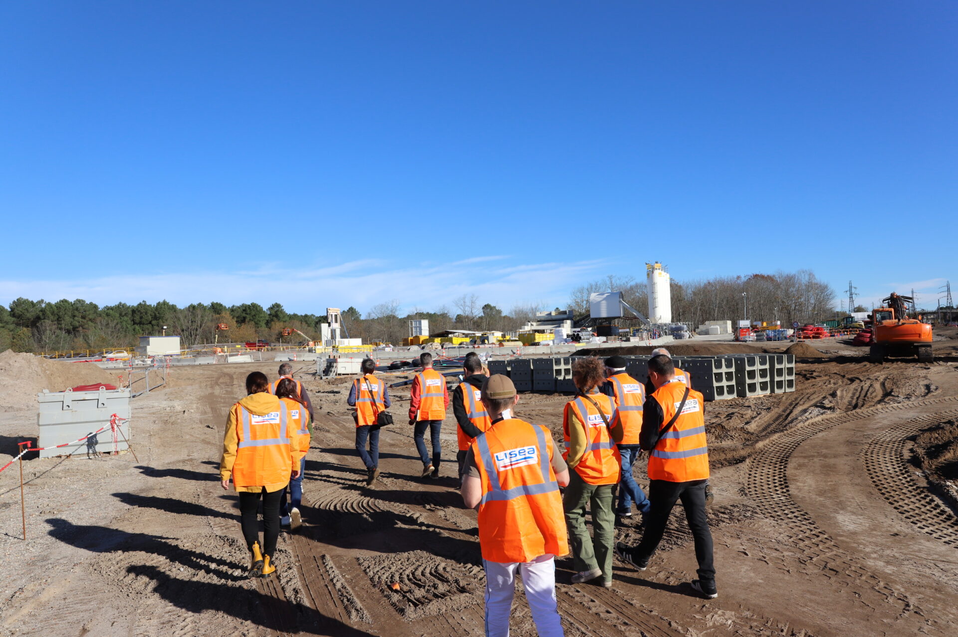 Un groupe de personnes en gilet orange marche sur un chantier. Au loin des batiments.