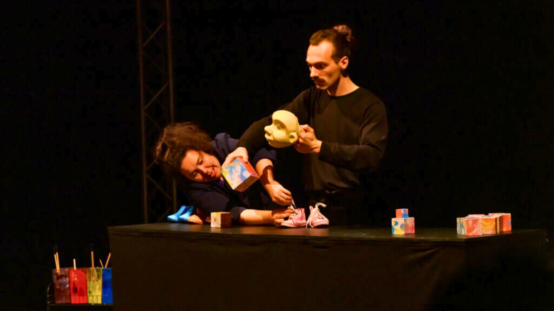 un homme et une femme sur scène habillés en noir qui tiennent des objets dans les mains. D'autres sont disposés sur une table -> spectacle qui sera présenté lors de la journée handicap et inclusion à mios