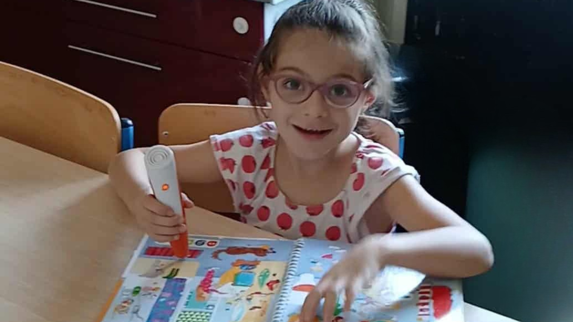 jeune fille de 6 ans habillée avec un tee-shirt blanc à pois rouge, elle porte des lunettes rose, elle est souriante et est assise à une table de cuisine avec un livre devant elle