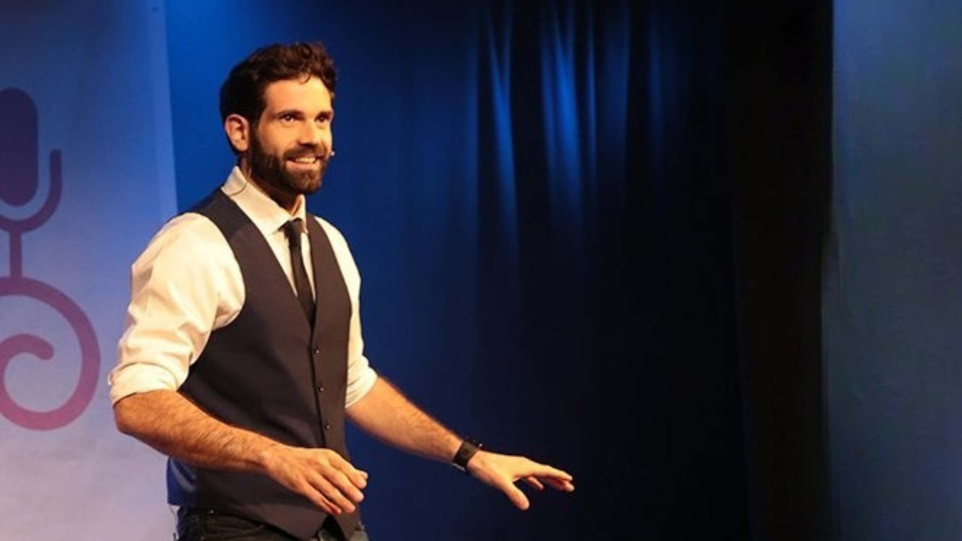 Marcus, humoriste : homme brun et barbu en chemise blanche avec une cravate noire et un gilet noir debout sur une scène avec un rideaux bleu foncé en arrière plan