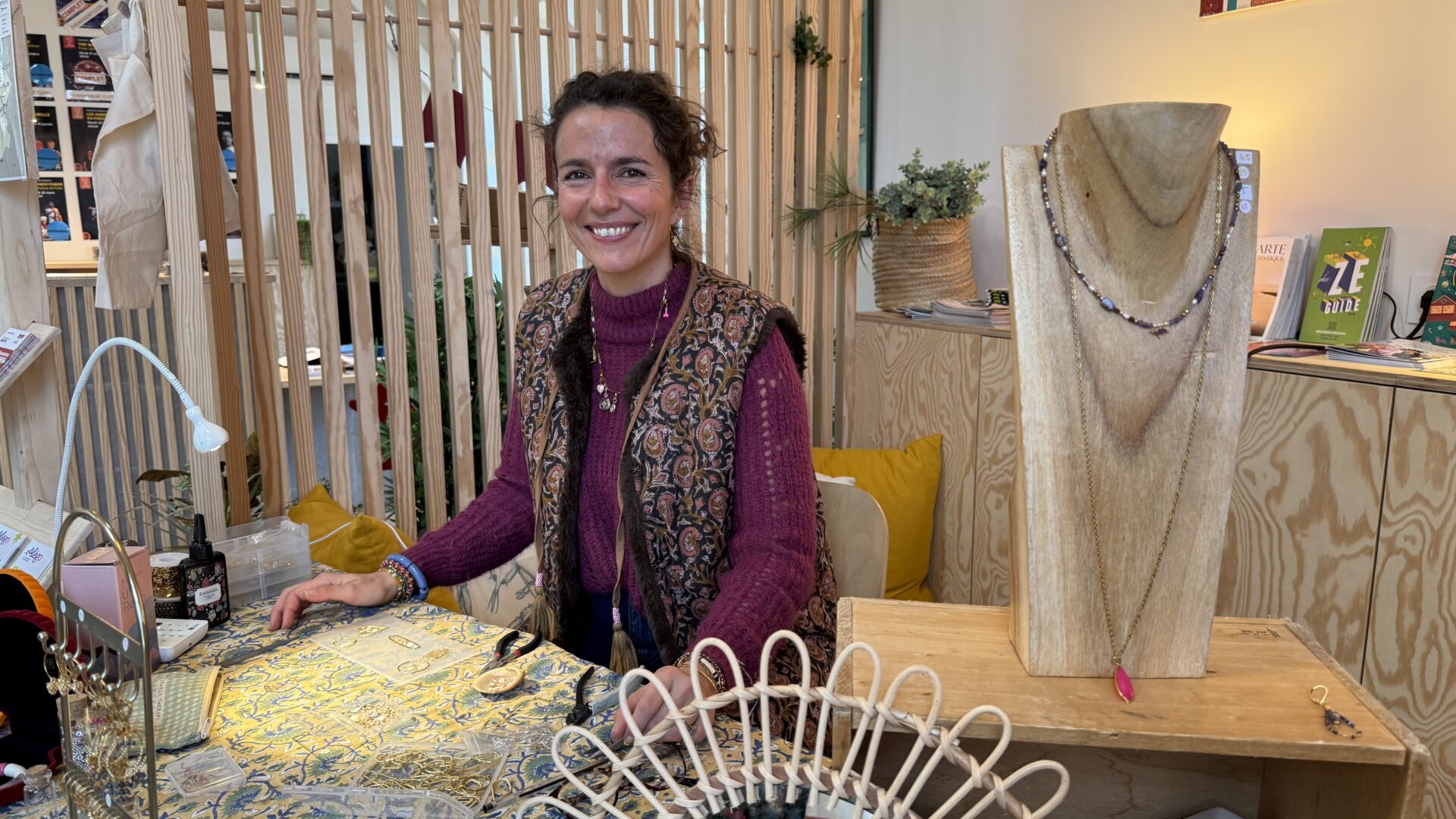 Une femme en pull violet et veston fleurit pose derrière un stand de bijoux