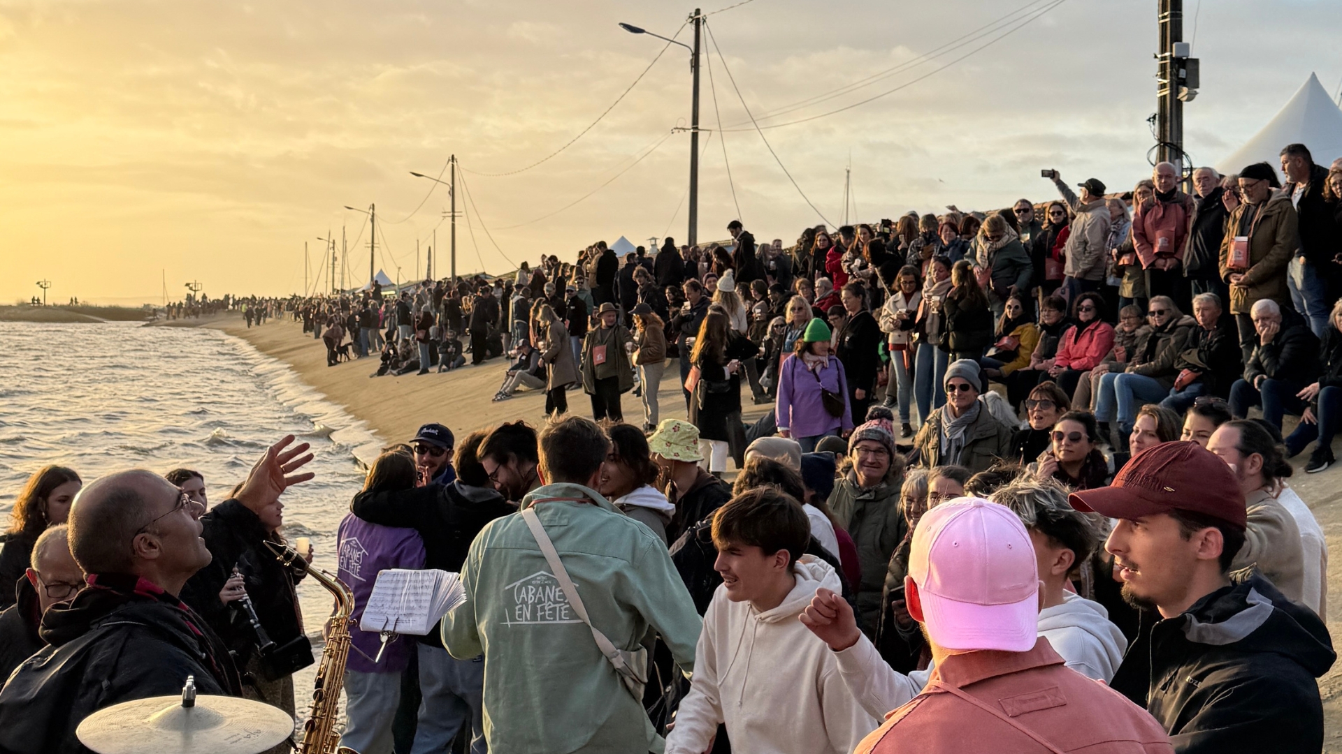 foule de personnes au port ostréicole d'andernos pour cabanes en fête 2025 avec la plage et le coucher de soleil en arrière plan
