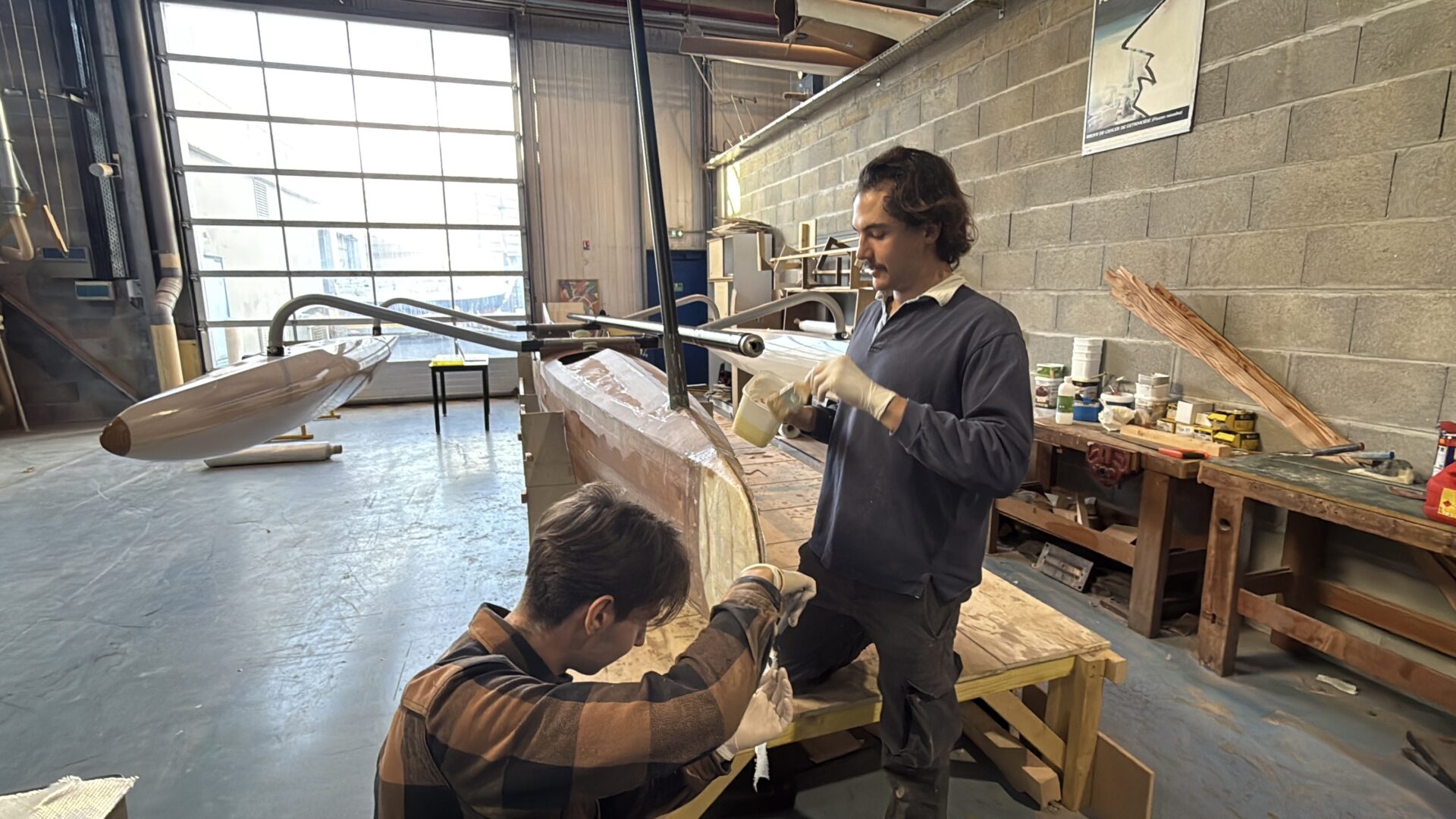 deux hommes construisent un trimaran dans un grand atelier éclairé par une grande baie-virée en arrière plan.