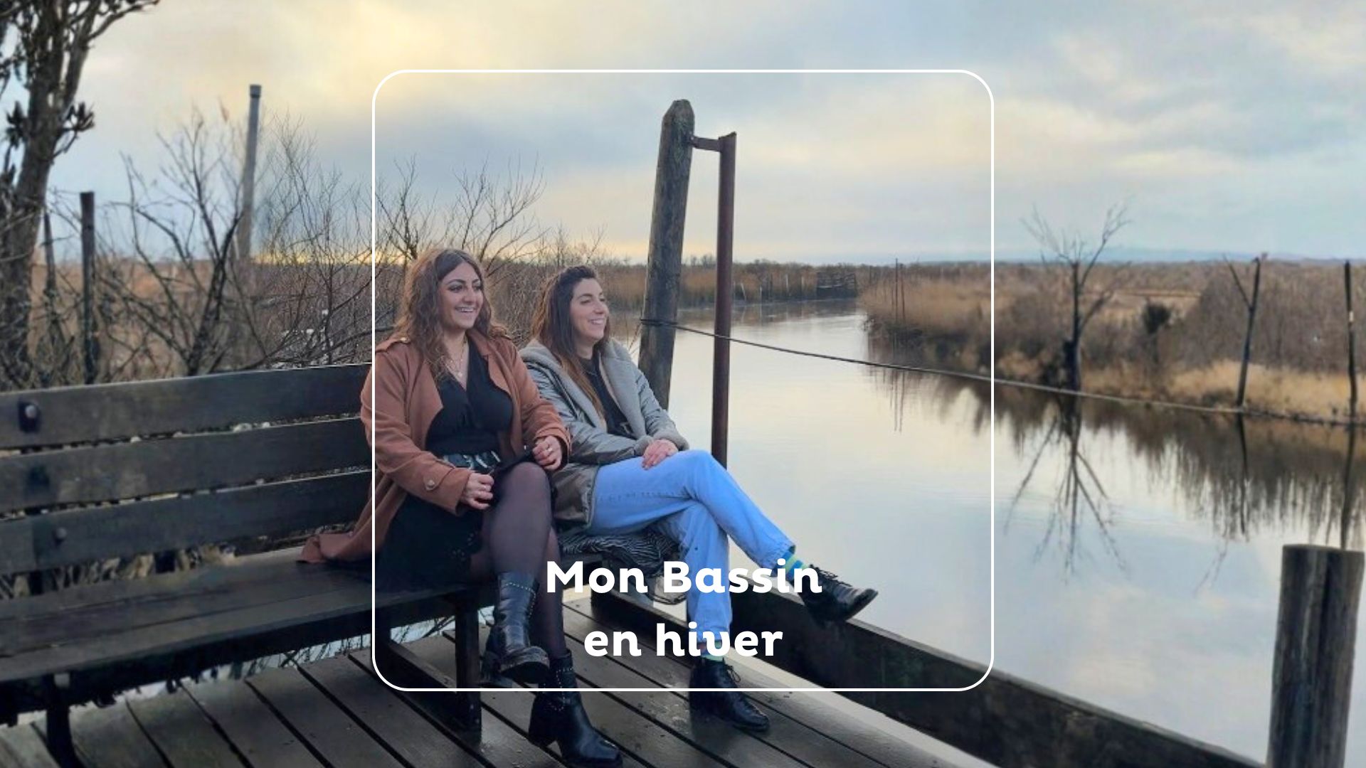 Deux femmes assises sur un banc en bois, souriantes, profitant d'une vue paisible sur un bassin en hiver. Le paysage est composé de végétation sèche et d'eau calme sous un ciel nuageux. Au centre, un encadré affiche le texte : « Mon Bassin en hiver ».