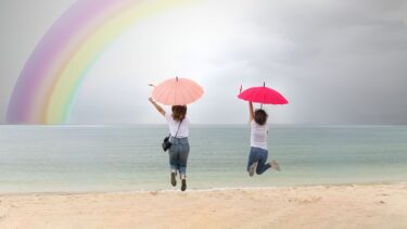 Deux femmes saute face à la mer avec de sparapluies colorés dans les Mains. En arrière plan ciel nuageux et arc-en-ciel