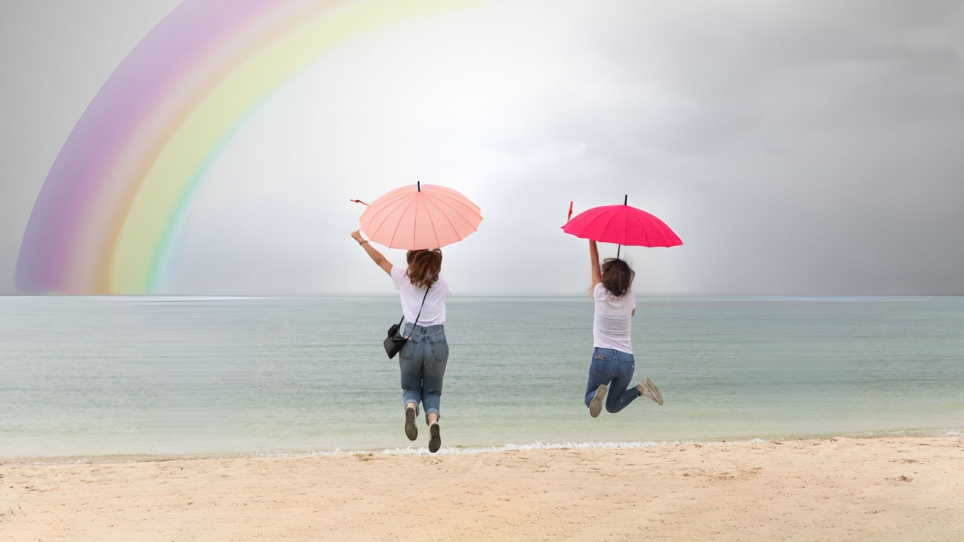 Deux femmes saute face à la mer avec de sparapluies colorés dans les Mains. En arrière plan ciel nuageux et arc-en-ciel