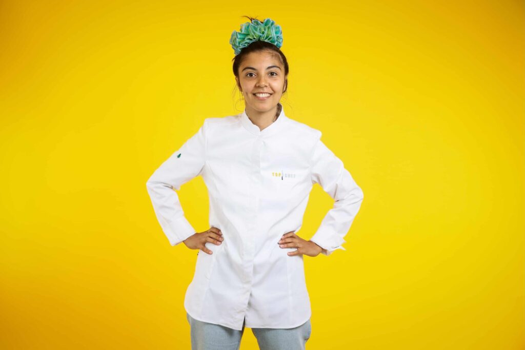 Une jeune femme en tenue de cuisine marquée Top Chef se tient devant un fond jaune. Elle a une coiffure verte dans les cheveux