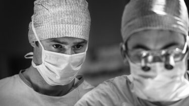 Photo en noir et blanc de deux hommes en blouse blanche de medecin, ils portent des masque et celui au premier plan porte des lunettes avec loupe.
