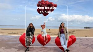 deux jeunes femmes posent devant la statue d'une petite fille assise sur un coeur et qui tient dans sa main de sballons coeurs rouoges. en arrière plan : la plage et la mer.