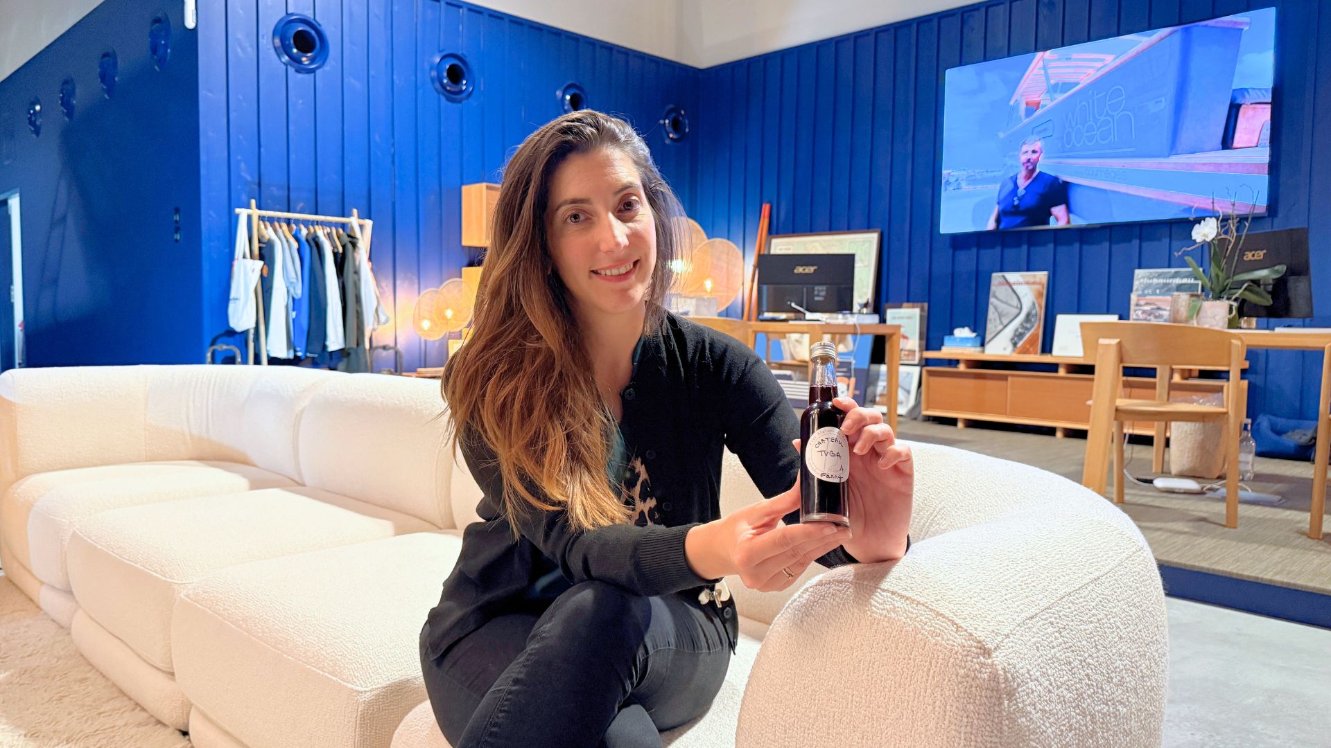 Une jeune femme aux cheveux long est assise dans un canapé blanc. Elle tient dans les mains une petite bouteille devin. derrière elle des bureaux et un mur en bois bleu.