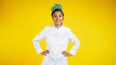 Une jeune femme en tenue de cuisine marquée Top Chef se tient devant un fond jaune. Elle a une coiffure verte dans les cheveux