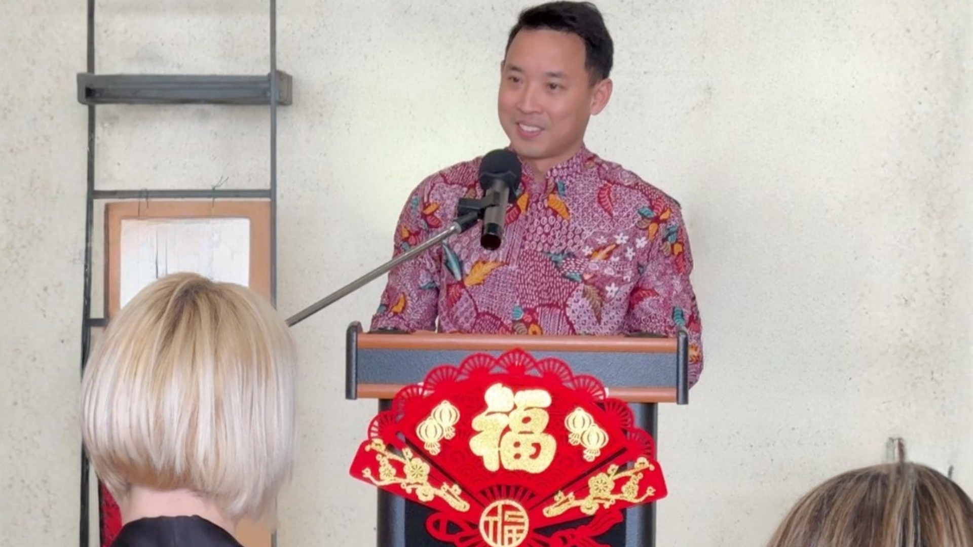 chef du détachement singapourien à Cazaux : homme souriant habillé d'une chemise colorée et placé derrière un pupitre avec un décor rouge et or. Des personnes de dos au premier rang