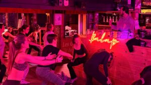 dans un bar, lumières rouges, des femmes qui font du pilates en tenue de sport. en arrière plan, un homme derrière les platines. Un néon avec l'inscription "balap" est apposé sur le bar en bois