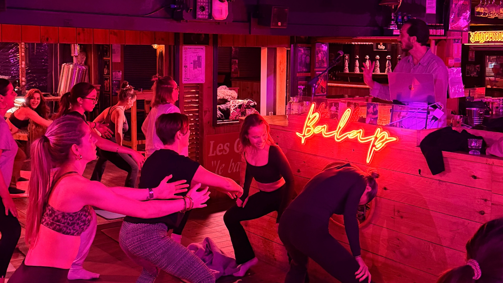 dans un bar, lumières rouges, des femmes qui font du pilates en tenue de sport. en arrière plan, un homme derrière les platines. Un néon avec l'inscription "balap" est apposé sur le bar en bois
