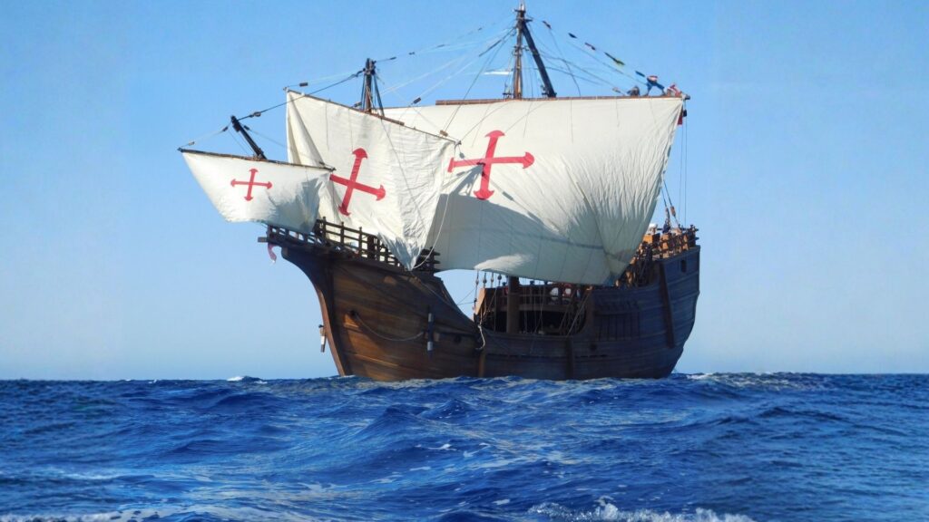 Un grand bateau en bois à 3 mats avec des voiles blanches marquées d'un symbole rouge