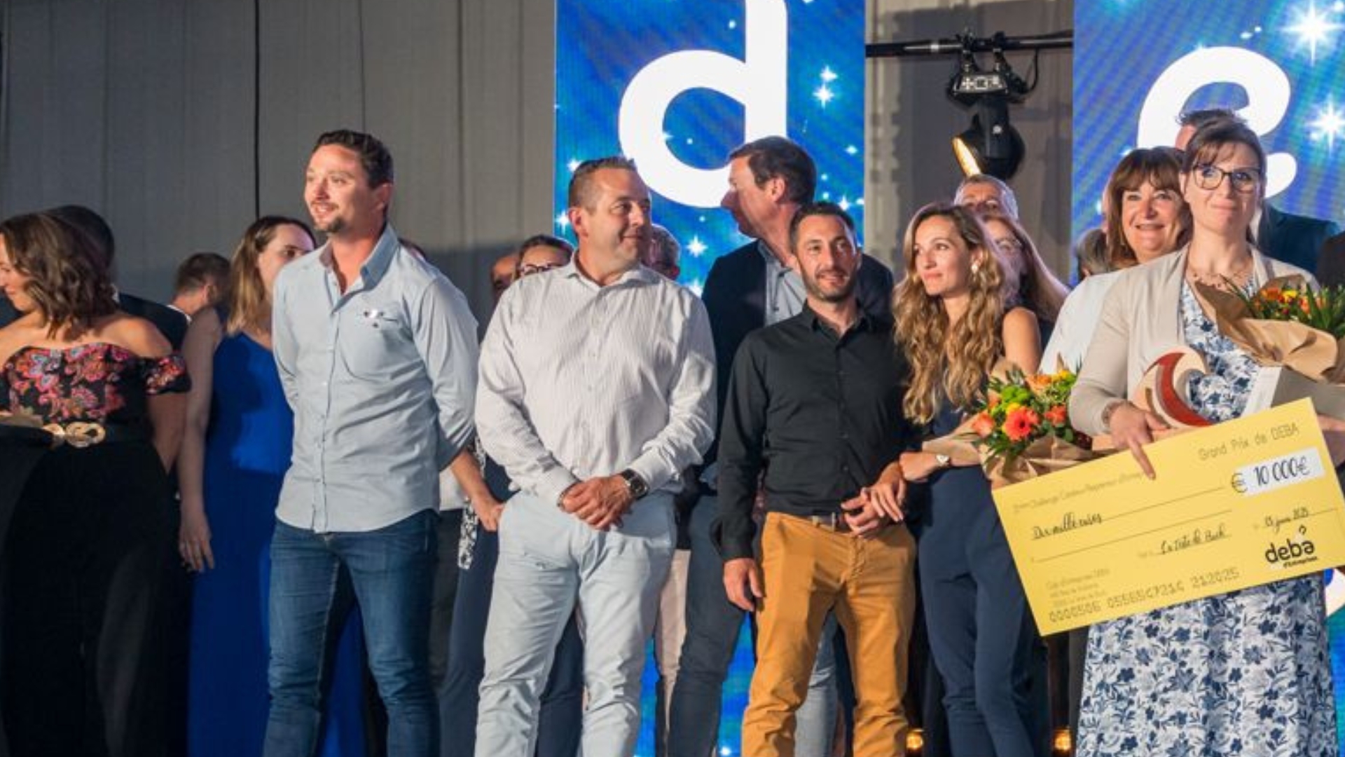 des personnes sur une scène. Un femme sur la droite tient un chèque en carton et un bouquet de fleurs