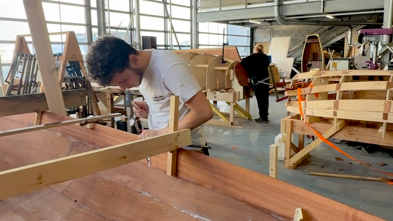 Un jeune homme en tee shirt blanc travaille le bois sur une coque de bateau dans un atelier éclairé par de grandes baies vitrées