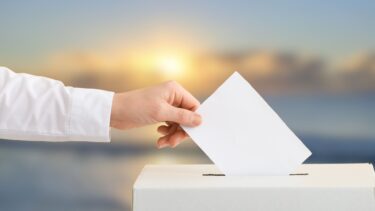 image d'illustration : une main tenant une enveloppe au dessus d'une urne de vote, fond flou d'une image de la plage avec un coucher de soleil