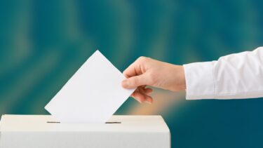 image d'illustration : une main tenant une enveloppe au dessus d'une urne de vote, fond flou d'une image aérienne du Bassin