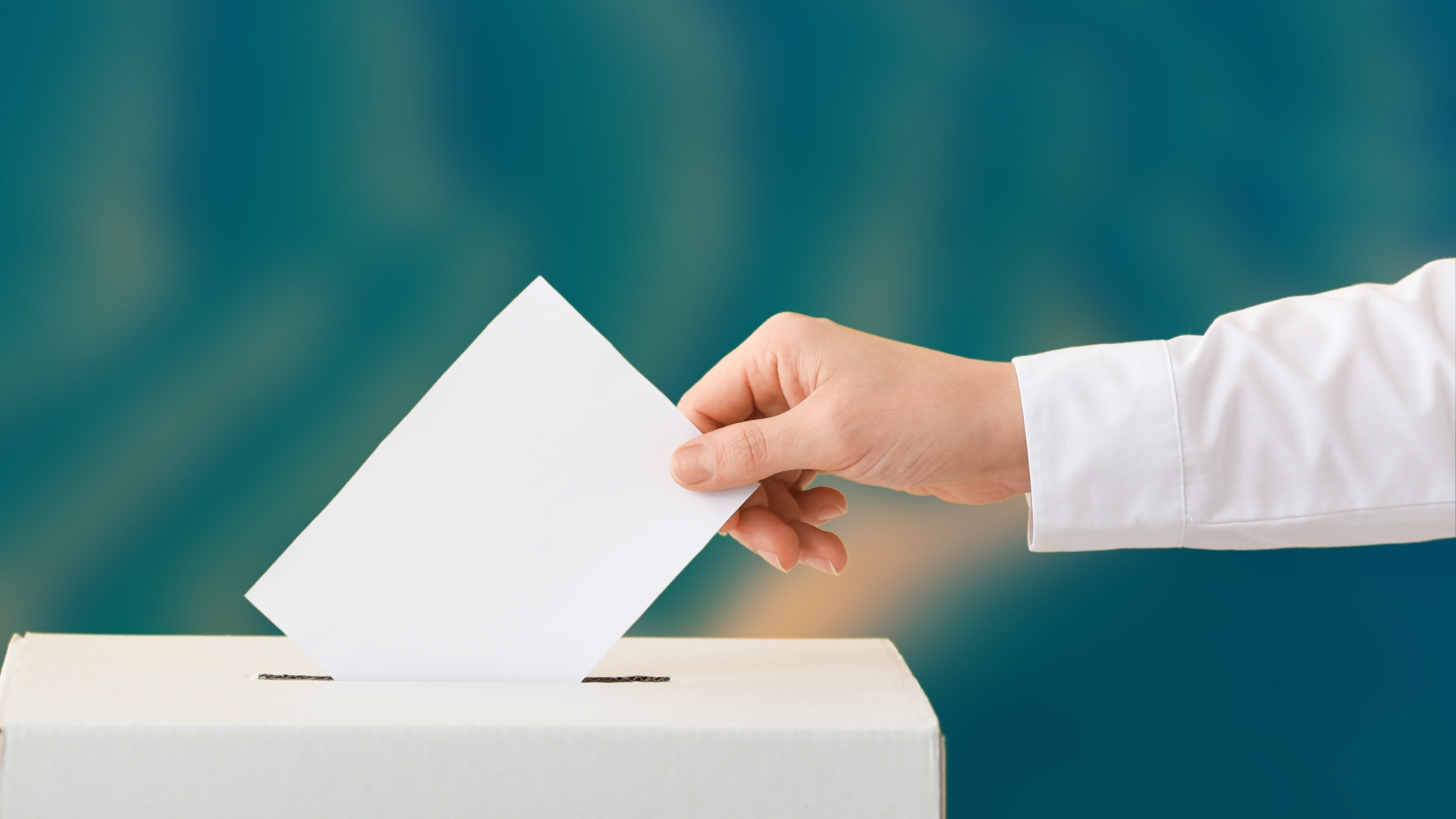 image d'illustration : une main tenant une enveloppe au dessus d'une urne de vote, fond flou d'une image aérienne du Bassin