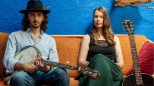 deux personnes assises sur un canapé orange. Un homme habillé d'une chemise bleu ciel et d'un chapeau sombre, il tient une guitare. Une femme avec une jupe verte et un débardeur noir. Arrière plan bleu