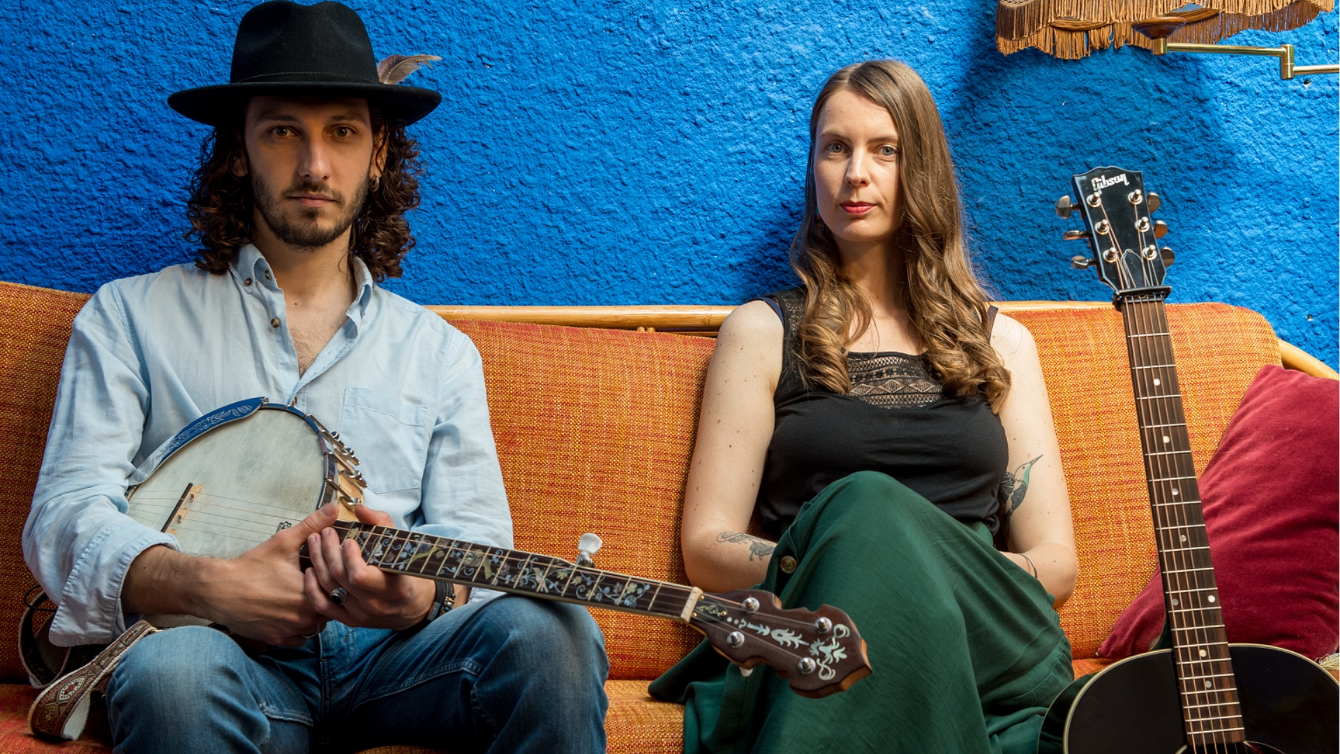 deux personnes assises sur un canapé orange. Un homme habillé d'une chemise bleu ciel et d'un chapeau sombre, il tient une guitare. Une femme avec une jupe verte et un débardeur noir. Arrière plan bleu