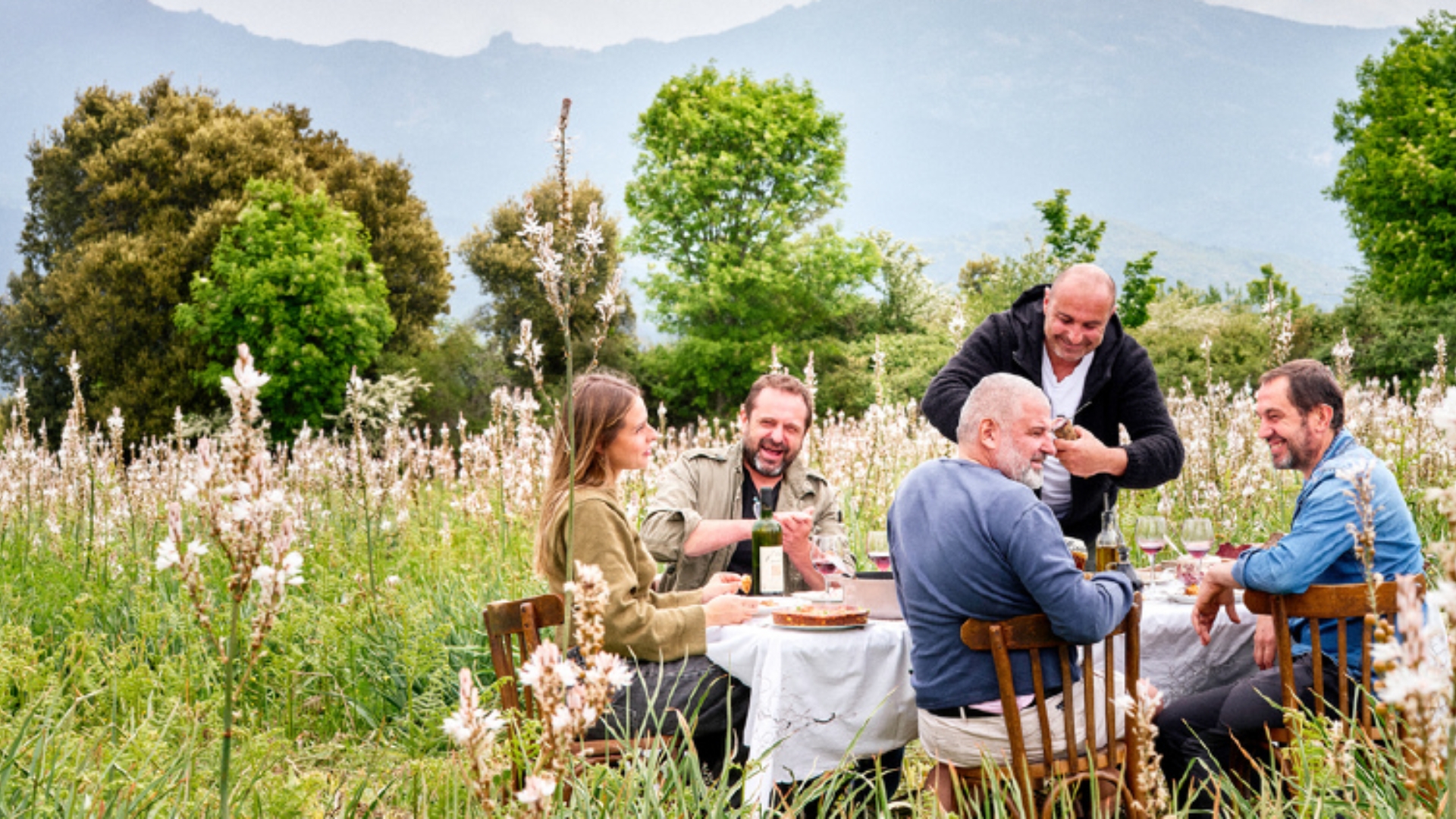 5 personnes assises à une table posées dans un champs de fleurs avec des montagnes en arrière-plan