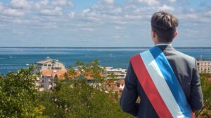 Une personne en costard et cheveux courts pose de dos avec une écharpe tricolore de maire. En arrière plan : une photo en hauteur de la ville d'Arcachon