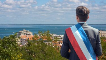 Une personne en costard et cheveux courts pose de dos avec une écharpe tricolore de maire. En arrière plan : une photo en hauteur de la ville d'Arcachon