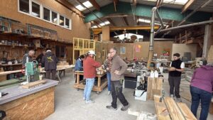 Un grand hangar en bois ouvert. `l'intérieur plusieurs personnes autour d'établit en train de bricoler.