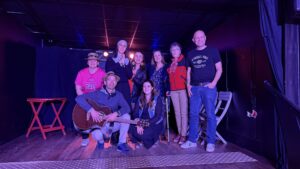 Un groupe de personne dont un homme tenant une guitare au premier plan prend la pose sur scène, éclairage rose.